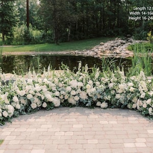 May include: A white floral archway with greenery, measuring 12 feet long, 14-16 inches wide, and 16-24 inches high. The archway is set up outdoors in front of a pond and a grassy area.