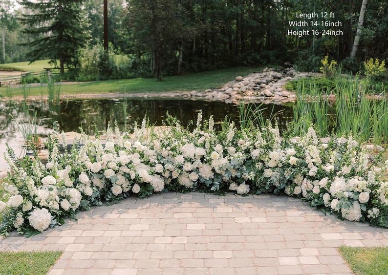 Op de afbeelding: Een witte bloemenboog met groen, met een lengte van 3,66 meter, een breedte van 35,56 tot 40,64 centimeter en een hoogte van 40,64 tot 61 centimeter. De boog is buiten opgesteld voor een vijver en een grasveld.