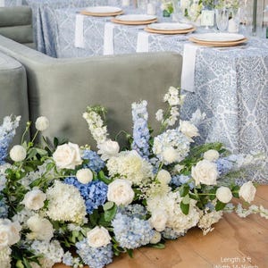 May include: A floral arrangement of white, light blue, and blue flowers, including roses and hydrangeas, adorns a wooden floor. The arrangement is in front of a gray sofa and tables set with white plates and patterned tablecloths. The text in the image says: Length 3 ft, Width 14-16 inch, Height 14-22 inch.