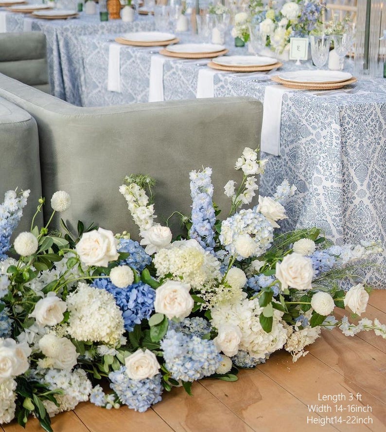 May include: A floral arrangement of white, light blue, and blue flowers, including roses and hydrangeas, adorns a wooden floor. The arrangement is in front of a gray sofa and tables set with white plates and patterned tablecloths. The text in the image says: Length 3 ft, Width 14-16 inch, Height 14-22 inch.
