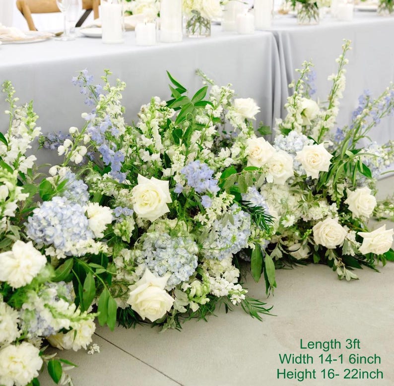 Puede incluir: Un pasillo de ceremonia de boda decorado con flores blancas y azules. Las flores est&aacute;n dispuestas en cascada, creando un ambiente rom&aacute;ntico y elegante. Las flores incluyen rosas blancas, hortensias azules y lisianthus blancos. El pasillo tiene aproximadamente 3 pies de largo, 14-16 pulgadas de ancho y 16-22 pulgadas de alto.