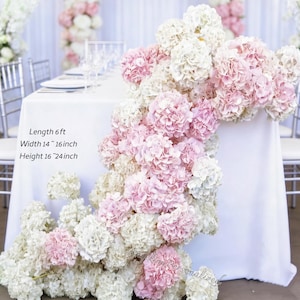 Könnte beinhalten: Ein weißes Tischtuch ist mit einer Kaskade aus rosa und weißen Hortensienblüten geschmückt. Der Tisch ist mit Tellern, Gläsern und Besteck gedeckt. Text auf dem Bild: Länge 1,8 m, Breite 36-41 cm, Höhe 41-61 cm. Der Tisch ist für eine Hochzeit oder eine besondere Veranstaltung gedeckt.
