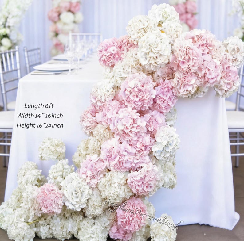 May include: A white tablecloth is decorated with cascading pink and white hydrangea flowers. The table is set with plates, glasses, and silverware. Text on the image reads: Length 6 ft, Width 14-16 inch, Height 16-24 inch. The table is set for a wedding or special event.