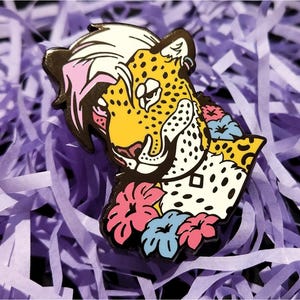 May include: A colorful enamel pin featuring a stylized leopard head with a yellow face, black spots, and pink and white hair. The leopard is adorned with pink, blue, and yellow flowers. The pin is set against a background of purple paper.