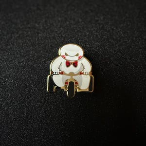 May include: A white and gold enamel pin featuring a cartoon character riding a tricycle. The character has a red bow tie and a stern expression. The tricycle has black wheels and a gold frame.