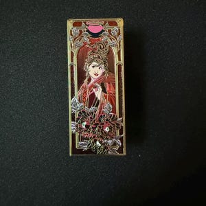 May include: A rectangular enamel pin featuring a woman in a red and gold design. The pin has a gold border with floral accents and the woman is holding a small object. The pin has the words "Lady Macbeth" at the bottom.