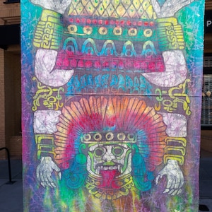 Puede incluir: Una pancarta de tela pintada a mano y colorida que representa una criatura mítica con un tocado de plumas, una boca grande y los brazos extendidos. La criatura está rodeada de intrincados patrones y diseños en tonos de azul, verde, rojo, amarillo y blanco.