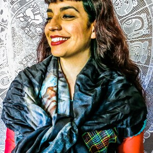May include: A woman wearing a dark scarf with a colourful plaid pattern and a blue and white design. The scarf is draped over a red top. The background features a white lace tapestry.