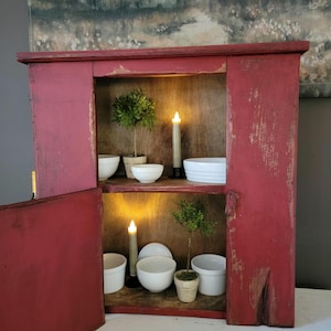 May include: A rustic, red-painted wooden cabinet with an open door, revealing two shelves. The shelves display white bowls, small potted plants, and lit candle-style lights. The cabinet has a distressed finish, adding to its vintage charm.