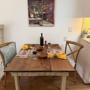 May include: A rustic wooden dining table set for two, featuring a bottle of wine, two glasses, and wooden bowls. The table is set with placemats and yellow napkins. A painting of a flower shop hangs on the wall above the table.