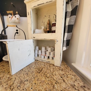 May include: A white cabinet with a distressed finish and a black and white checkered towel hanging on the side. The cabinet has two shelves, the top shelf has a small white ceramic deer figurine, a small pine tree, and a jar of jam. The bottom shelf has a collection of single-serve coffee pods.