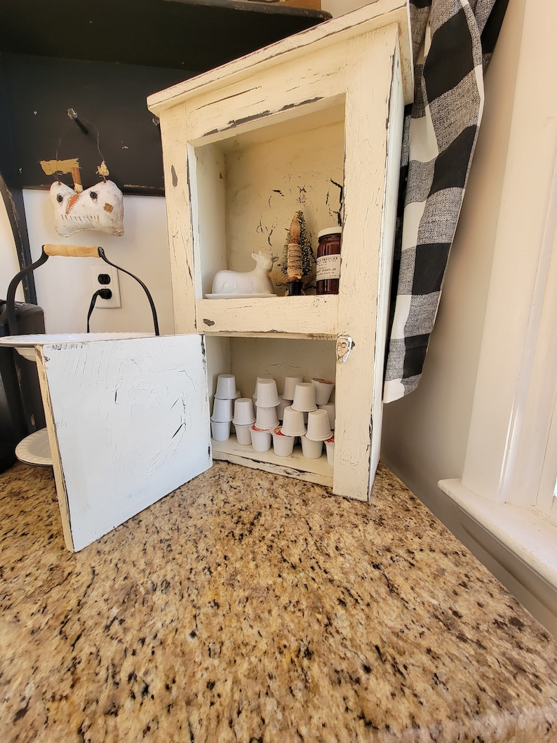 May include: A white cabinet with a distressed finish and a black and white checkered towel hanging on the side. The cabinet has two shelves, the top shelf has a small white ceramic deer figurine, a small pine tree, and a jar of jam. The bottom shelf has a collection of single-serve coffee pods.