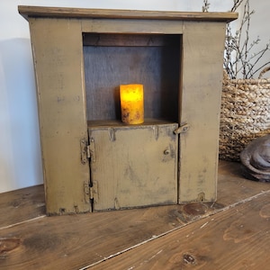 May include: A distressed, olive green wooden cabinet with a small door and a shelf. Inside the cabinet is a lit, yellow candle. The cabinet is sitting on a wooden table.