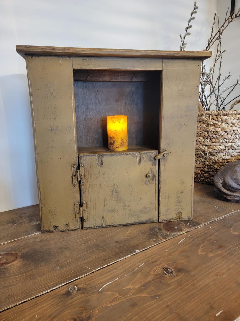 May include: A distressed, olive green wooden cabinet with a small door and a shelf. Inside the cabinet is a lit, yellow candle. The cabinet is sitting on a wooden table.