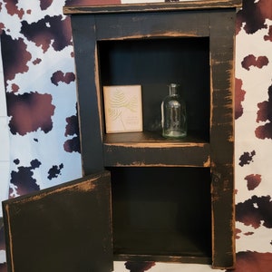 May include: A black wooden cabinet with a distressed finish. The cabinet has two shelves and a door that opens to reveal an empty shelf. The cabinet is sitting on a white and brown cowhide rug. A small sign with the text "So much makes me happy" is on the top shelf. A clear glass bottle is on the top shelf.