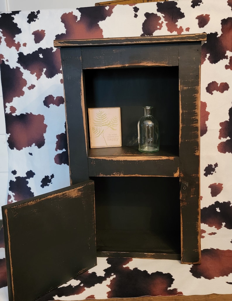 May include: A black wooden cabinet with a distressed finish. The cabinet has two shelves and a door that opens to reveal an empty shelf. The cabinet is sitting on a white and brown cowhide rug. A small sign with the text "So much makes me happy" is on the top shelf. A clear glass bottle is on the top shelf.