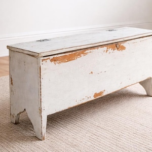 May include: A white, rectangular wooden chest with a hinged lid. The chest has a distressed, aged appearance with visible wear and tear, revealing the underlying wood. It stands on four angled legs, and the overall design suggests a vintage or antique style.