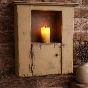 May include: A distressed, mustard-yellow wall cabinet with a small door and a lit candle inside. The cabinet is mounted on a brick wall. A vase of lavender sits on a wooden surface.