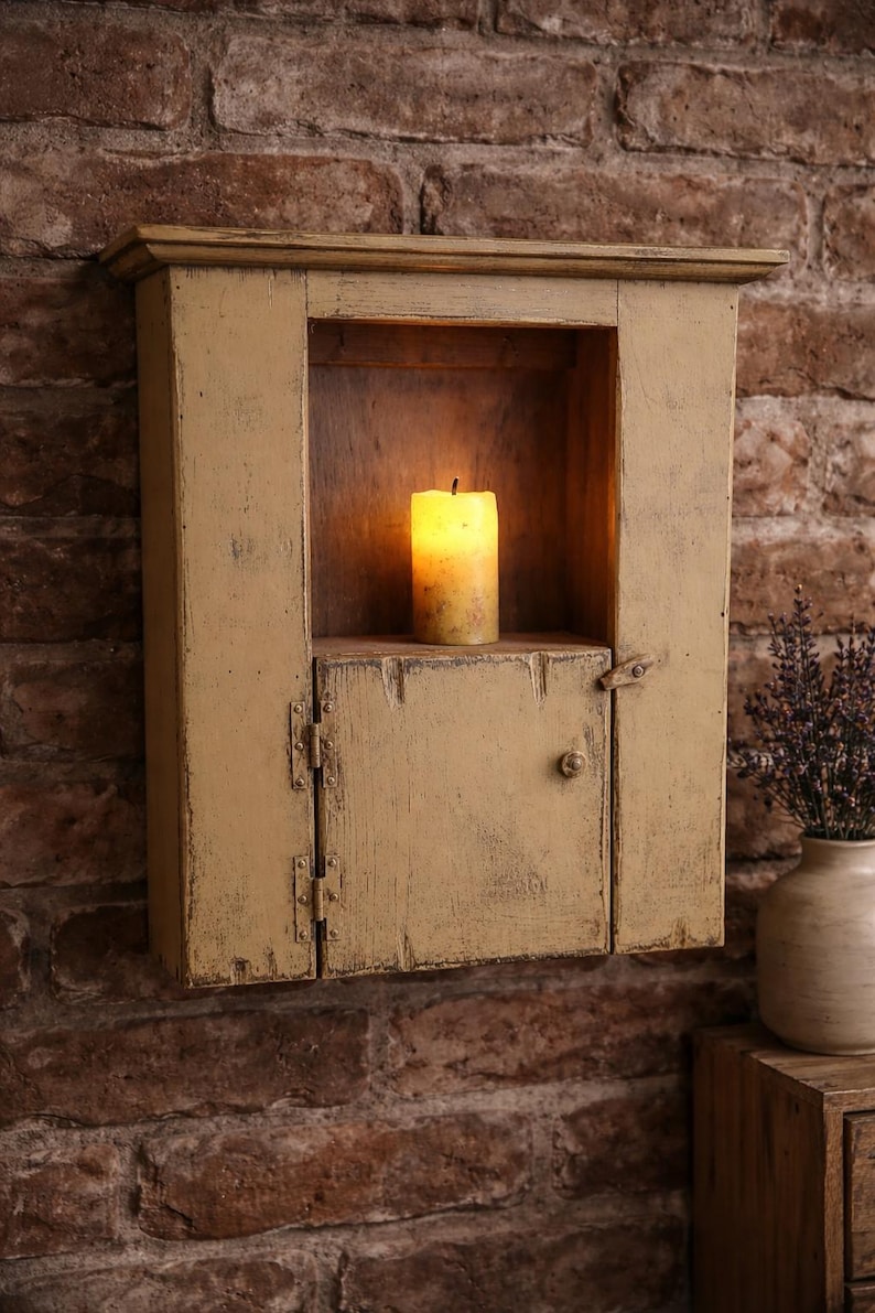 May include: A distressed, mustard-yellow wall cabinet with a small door and a lit candle inside. The cabinet is mounted on a brick wall. A vase of lavender sits on a wooden surface.