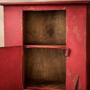 May include: A distressed red wooden cabinet with two doors open, revealing a two-shelf interior. The cabinet has a rustic, aged appearance with visible wood grain and paint chipping, suggesting a vintage or farmhouse style.