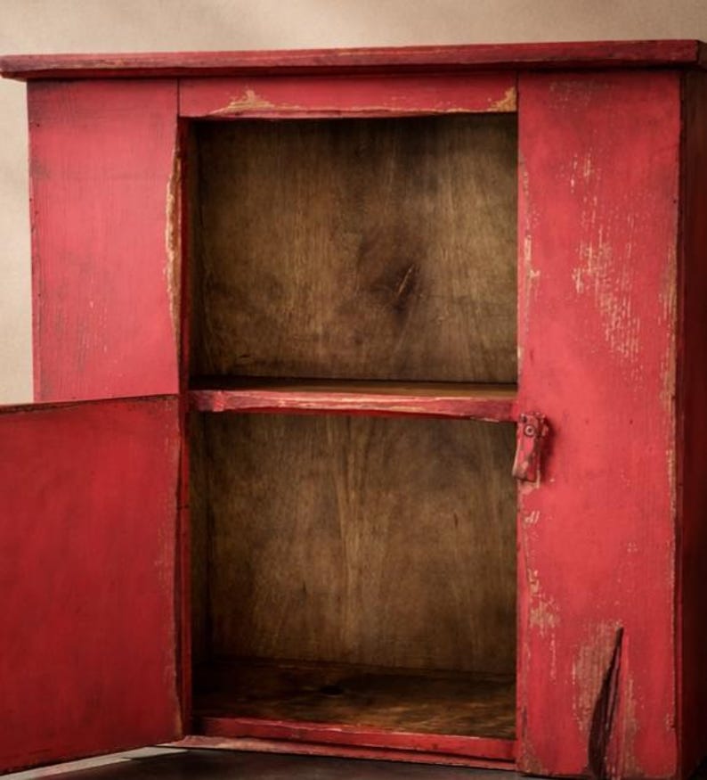 May include: A distressed red wooden cabinet with two doors open, revealing a two-shelf interior. The cabinet has a rustic, aged appearance with visible wood grain and paint chipping, suggesting a vintage or farmhouse style.