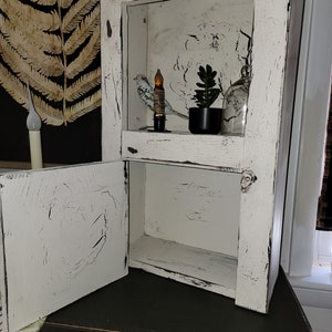 May include: A white wooden cabinet with a distressed finish and a single door that is open. The cabinet has two shelves, and the top shelf is decorated with a small bird figurine, a candle, and a potted succulent plant.