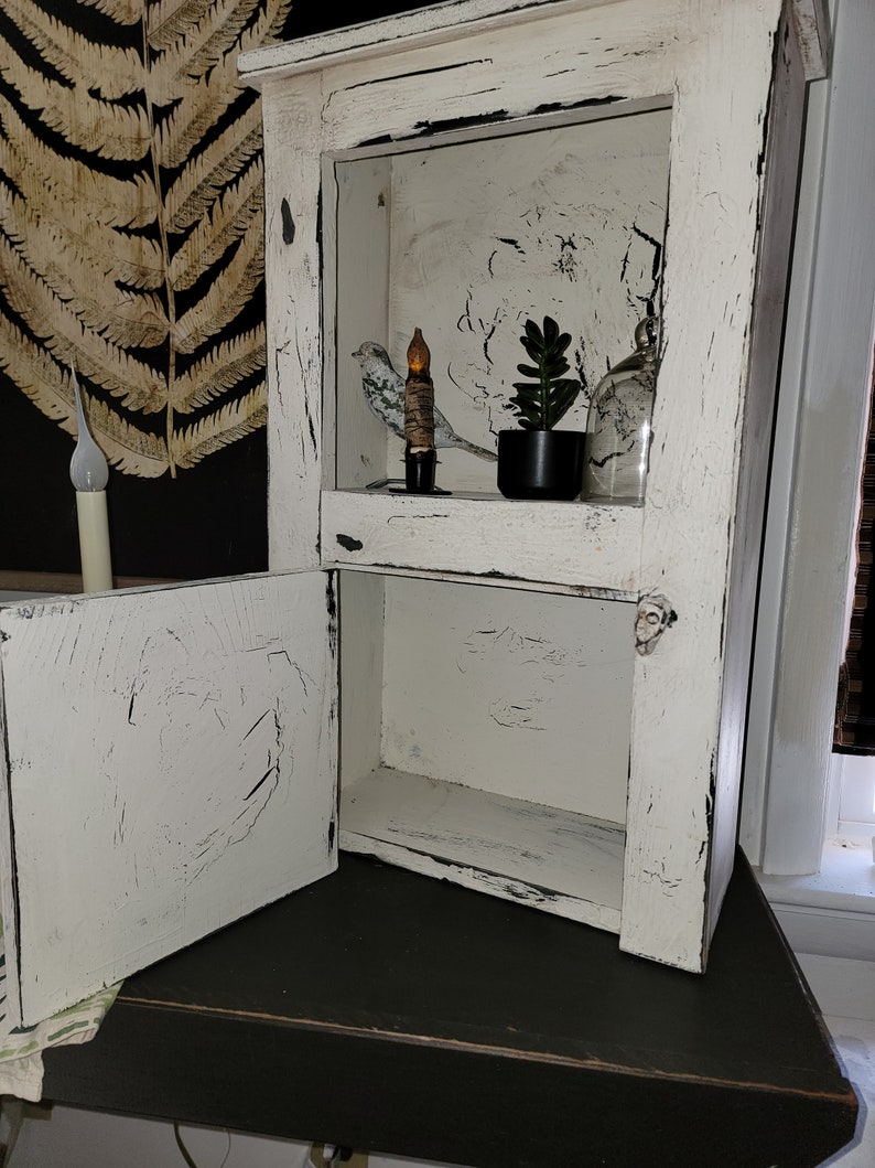 May include: A white wooden cabinet with a distressed finish and a single door that is open. The cabinet has two shelves, and the top shelf is decorated with a small bird figurine, a candle, and a potted succulent plant.