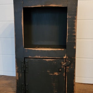 May include: A black wooden cabinet with a distressed finish. It has a single door with hinges and a small shelf above the door. The cabinet is about 30 inches tall.