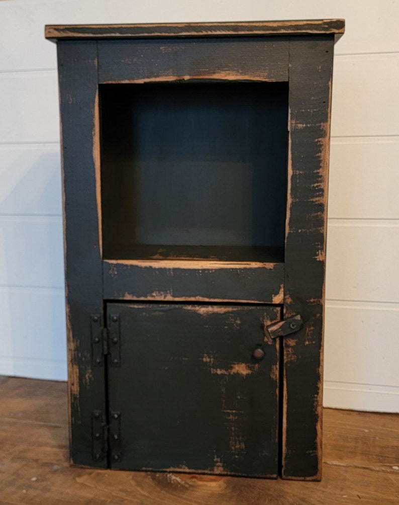 May include: A black wooden cabinet with a distressed finish. It has a single door with hinges and a small shelf above the door. The cabinet is about 30 inches tall.