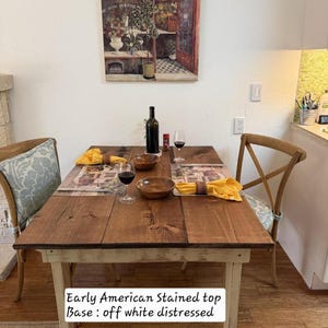 May include: A rectangular dining table with an Early American stained wood top and an off-white distressed base. The table is set with two wooden bowls, two wine glasses filled with red wine, a wine bottle, and two yellow napkins with rings.
