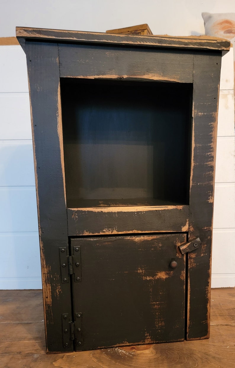 May include: A black wooden cabinet with a distressed finish. It has a single door with a metal hinge and a small shelf inside. The cabinet is about 30 inches tall.