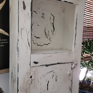 May include: White wooden cabinet with a distressed finish and black accents. The cabinet has a shelf and a door with hinges.