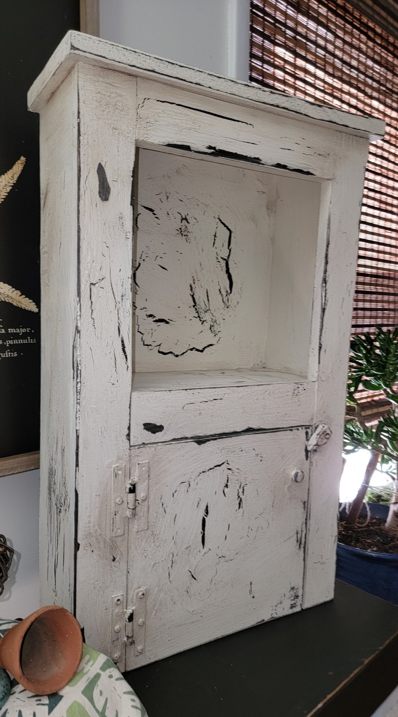 May include: White wooden cabinet with a distressed finish and black accents. The cabinet has a shelf and a door with hinges.