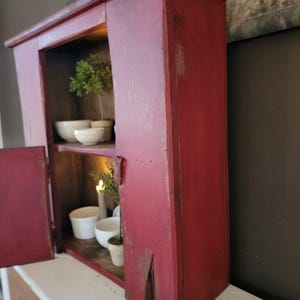 May include: A rustic, red-painted wooden cabinet with a distressed finish. The cabinet has two shelves, and the door is open, revealing white bowls and a small potted plant. The cabinet is sitting on a white surface.