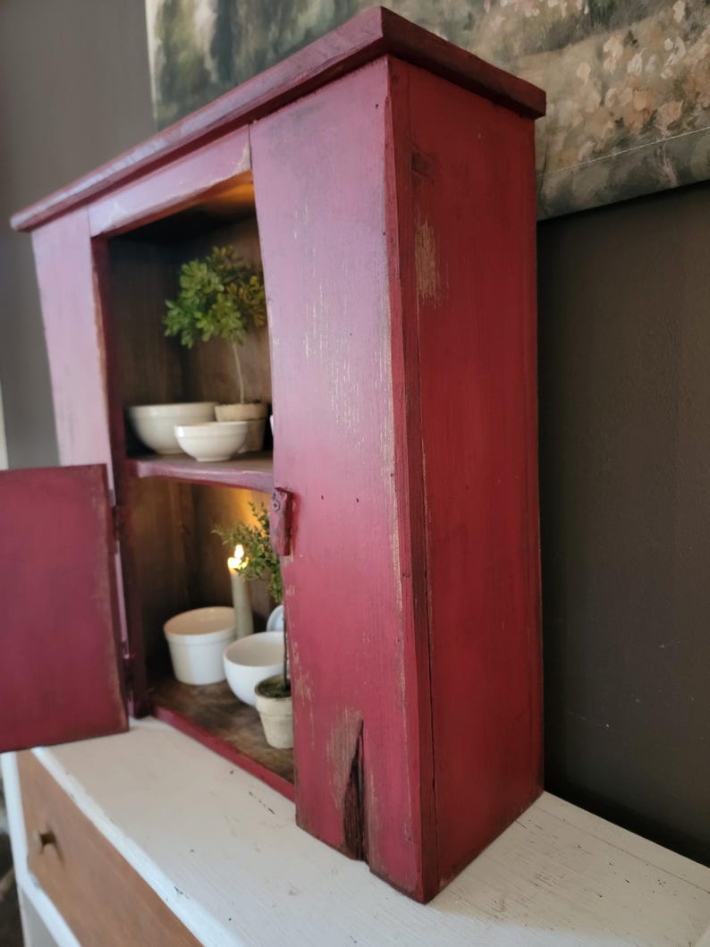 May include: A rustic, red-painted wooden cabinet with a distressed finish. The cabinet has two shelves, and the door is open, revealing white bowls and a small potted plant. The cabinet is sitting on a white surface.