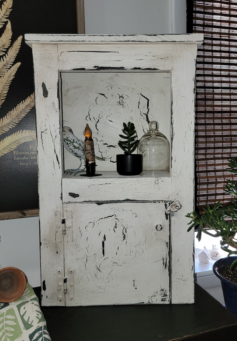May include: A white distressed wooden cabinet with a glass door and a shelf inside. The shelf has a small potted succulent plant, a glass cloche, and a candle. The cabinet has a rustic farmhouse style.