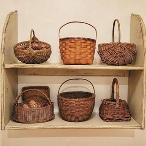 May include: A display of six woven baskets of various shapes and sizes on a two-tiered, off-white shelf. The baskets are made of natural brown materials and feature handles. The shelf is mounted on a light-colored wall.