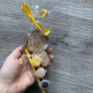 Pu&ograve; includere: Un kit di artigianato in legno con un cane dei cartoni animati che indossa un cappello da pompiere. Il kit include tre piccoli barattoli di vernice gialla, beige e rossa, e un pennello giallo.