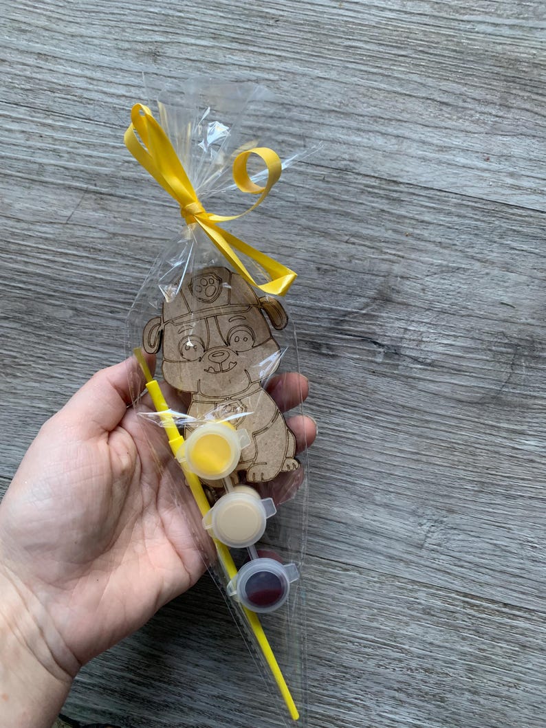 Pu&ograve; includere: Un kit di artigianato in legno con un cane dei cartoni animati che indossa un cappello da pompiere. Il kit include tre piccoli barattoli di vernice gialla, beige e rossa, e un pennello giallo.