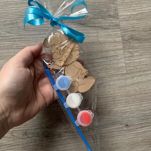 Pu&ograve; includere: Un kit artigianale in legno a forma di Puffo in un sacchetto di cellophane trasparente, legato con un nastro blu brillante. Il kit include tre vasetti di vernice piccoli in blu, bianco e rosso e un pennello blu. Il Puffo &egrave; non verniciato.