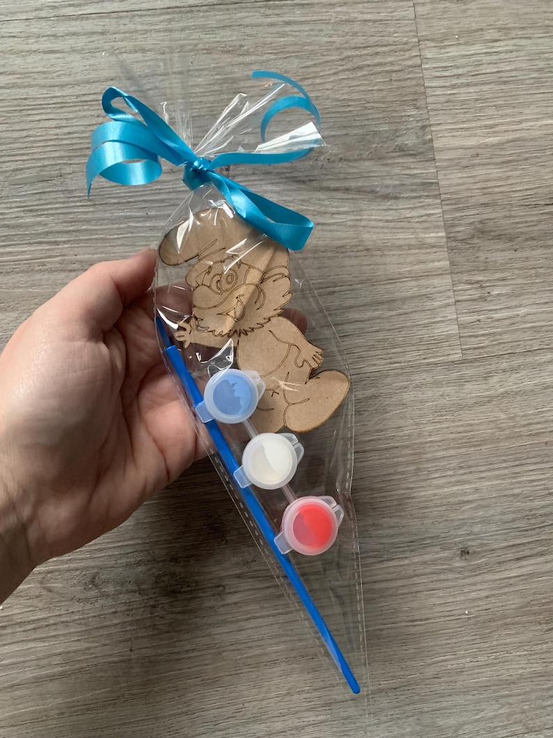 Pu&ograve; includere: Un kit artigianale in legno a forma di Puffo in un sacchetto di cellophane trasparente, legato con un nastro blu brillante. Il kit include tre vasetti di vernice piccoli in blu, bianco e rosso e un pennello blu. Il Puffo &egrave; non verniciato.