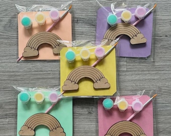 May include: Five craft kits in clear plastic packaging, each containing a wooden rainbow cutout, a small paintbrush, and three miniature paint pots in teal, yellow, and pink, mounted on square cardstock in peach, purple, yellow, mint, and pink.