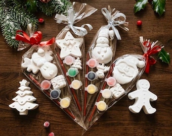 May include: Three clear plastic cone-shaped bags containing white plaster holiday figures, including a reindeer, star, snowman, and Santa Claus, each paired with a small paintbrush and a strip of five paint pots, tied with red or white ribbons.