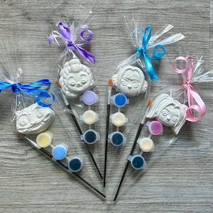 Puede incluir: Cuatro kits de pintura para hacer uno mismo, cada uno en una bolsa de celofán transparente atada con una cinta de colores. Cada kit incluye un personaje de yeso blanco, un pincel pequeño y una paleta de pintura con varios colores. Los personajes incluyen un gato de Cheshire y tres caras femeninas.