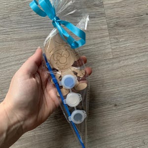 Pu&ograve; includere: Un sacchetto di cellophane trasparente legato con un nastro azzurro contiene un kit artigianale in legno a forma di Puffo. Il kit include un ritaglio di legno, tre vasetti di vernice piccoli con vernice blu, bianca e grigia e un pennello blu.