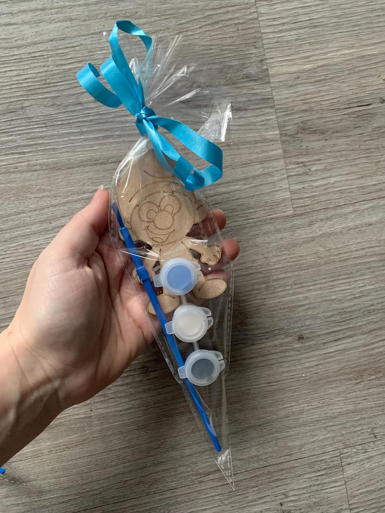 Pu&ograve; includere: Un sacchetto di cellophane trasparente legato con un nastro azzurro contiene un kit artigianale in legno a forma di Puffo. Il kit include un ritaglio di legno, tre vasetti di vernice piccoli con vernice blu, bianca e grigia e un pennello blu.