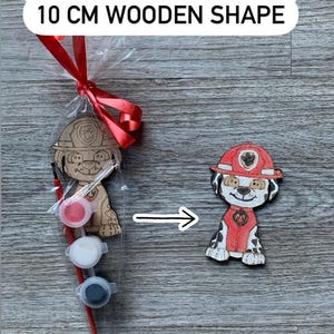 Pu&ograve; includere: Un kit di pittura in legno per dipingere il tuo personaggio con un cane dei cartoni animati che indossa un cappello da pompiere rosso. Il kit include tre piccoli barattoli di vernice, un pennello e una forma in legno di 10 cm. "NEW WOODEN OPTION! 10 CM WOODEN SHAPE" &egrave; scritto sulla confezione.