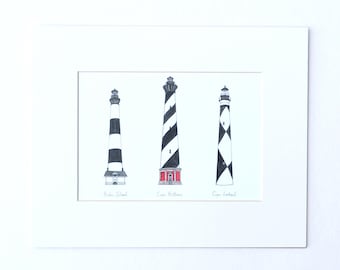 May include: A framed print featuring three lighthouse illustrations. The lighthouses are depicted in black and white with varying stripe patterns. The middle lighthouse has a red base. The names of the lighthouses are written below them.