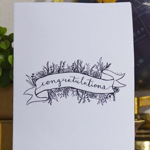 May include: A white card with a black and white illustration of a banner with the word "congratulations" written on it. The banner is surrounded by a wreath of flowers and leaves.