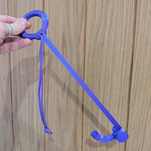 May include: A purple 3D-printed reacher tool featuring a circular handle at one end and a curved hook at the other, connected by a long, straight arm. A thin purple cord is attached to the handle, providing a loop for easy gripping and storage.